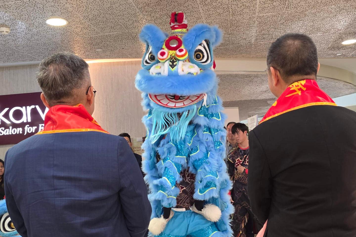 Two persons wearing red shawls are facing a light blue dragon performer at the opening ceremony of the Zeiss Vision Center in Singapore