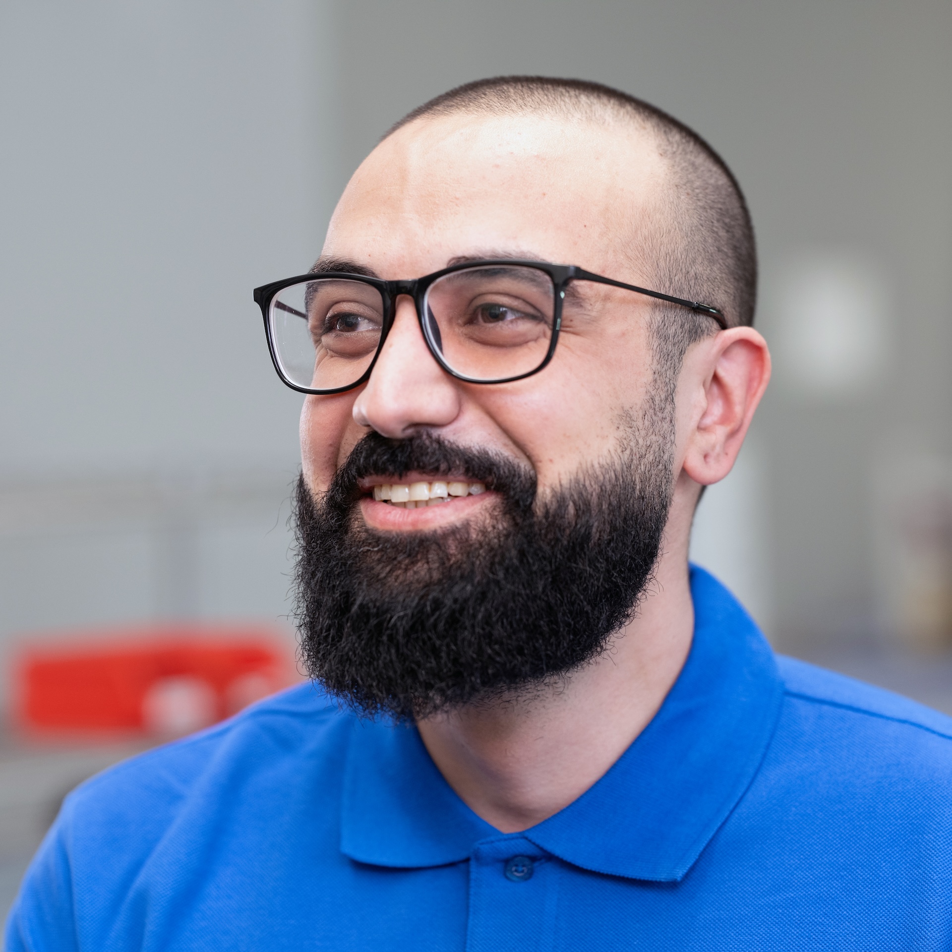 Ali El-Toufaili, a bearded man wearing glasses and a blue polo shirt is smiling in a bright indoor setting.