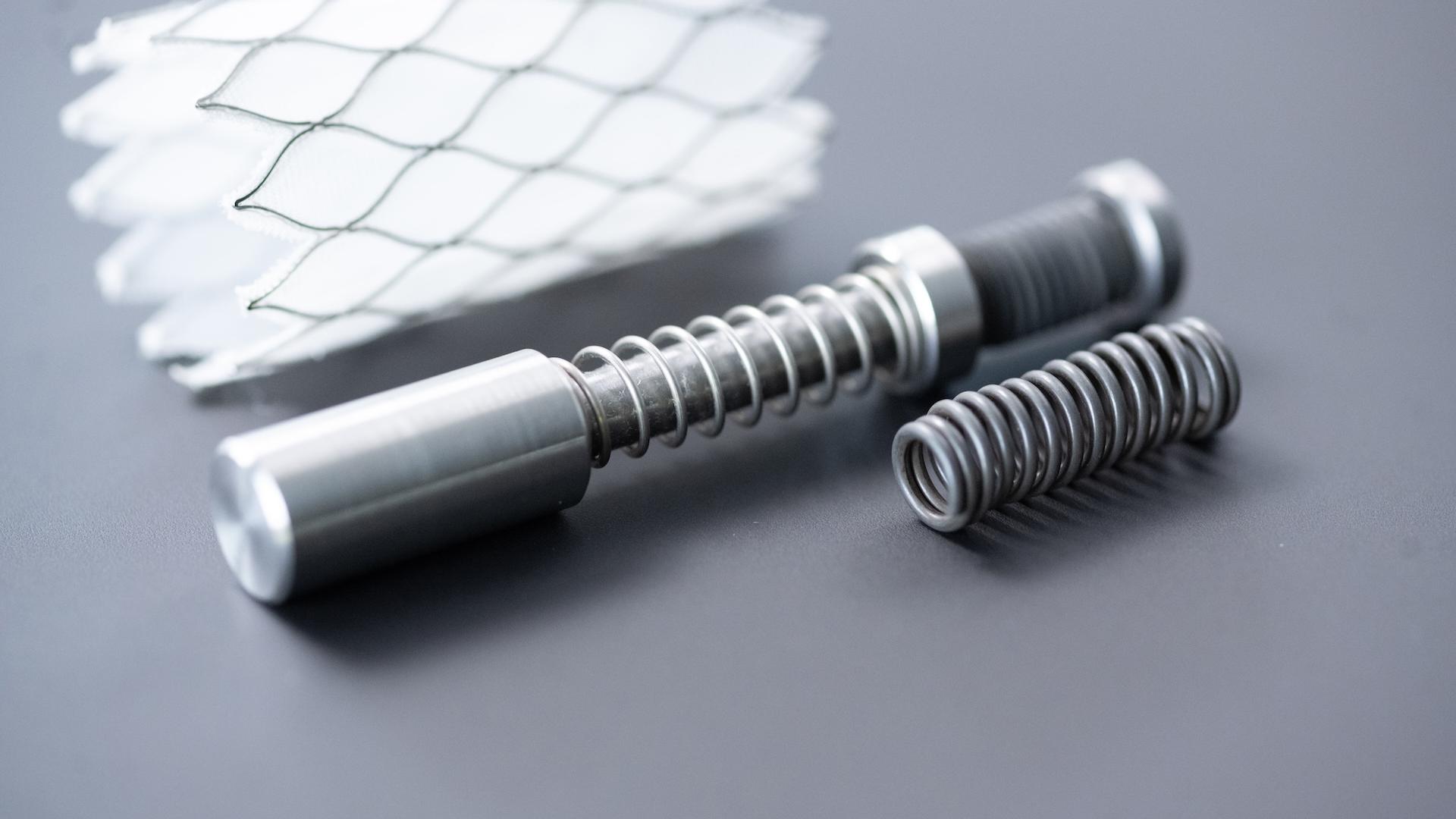 Close-up of precision-engineered metal springs and components alongside a white mesh structure on a grey surface.