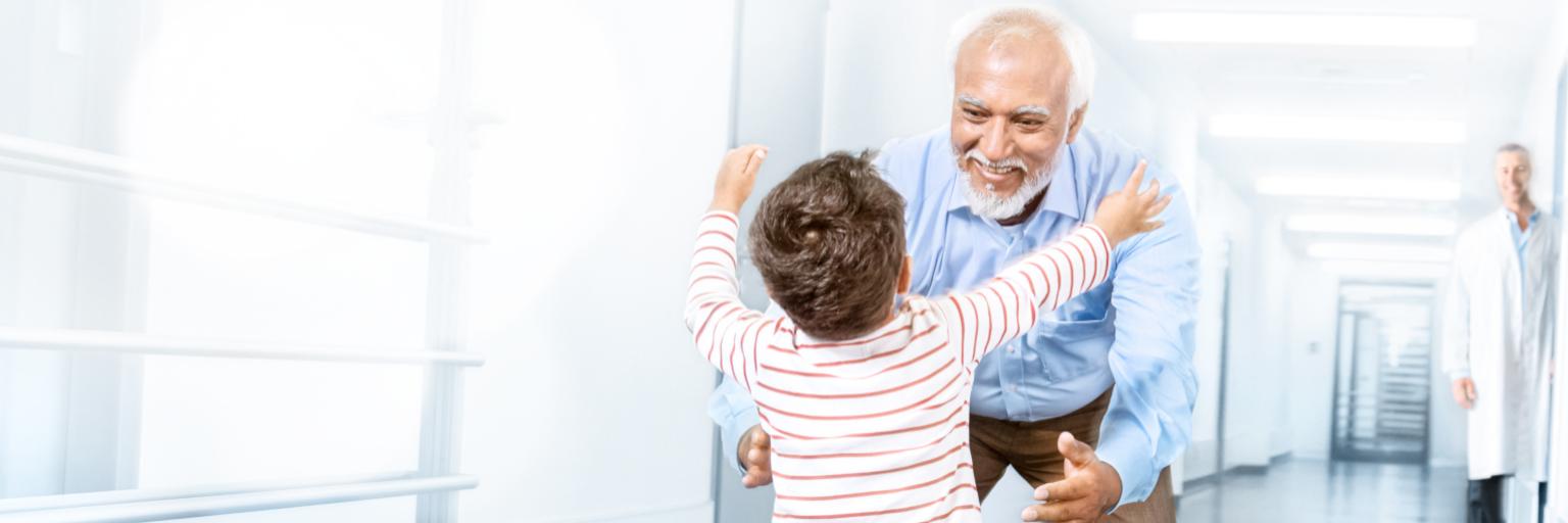 An elderly man warmly embraces a young child in a bright, modern hallway, with a ZEISS medical professional observing in the background.