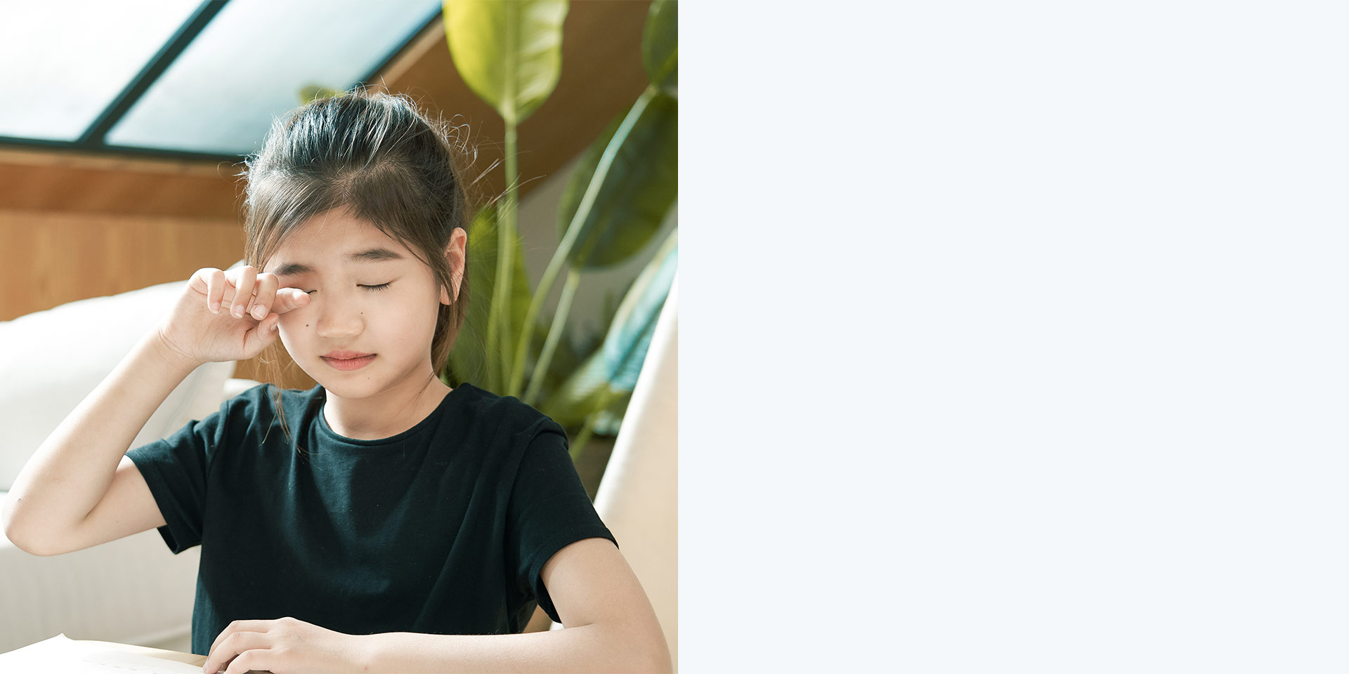 Young girl without glasses rubbing her eyes while reading a book.