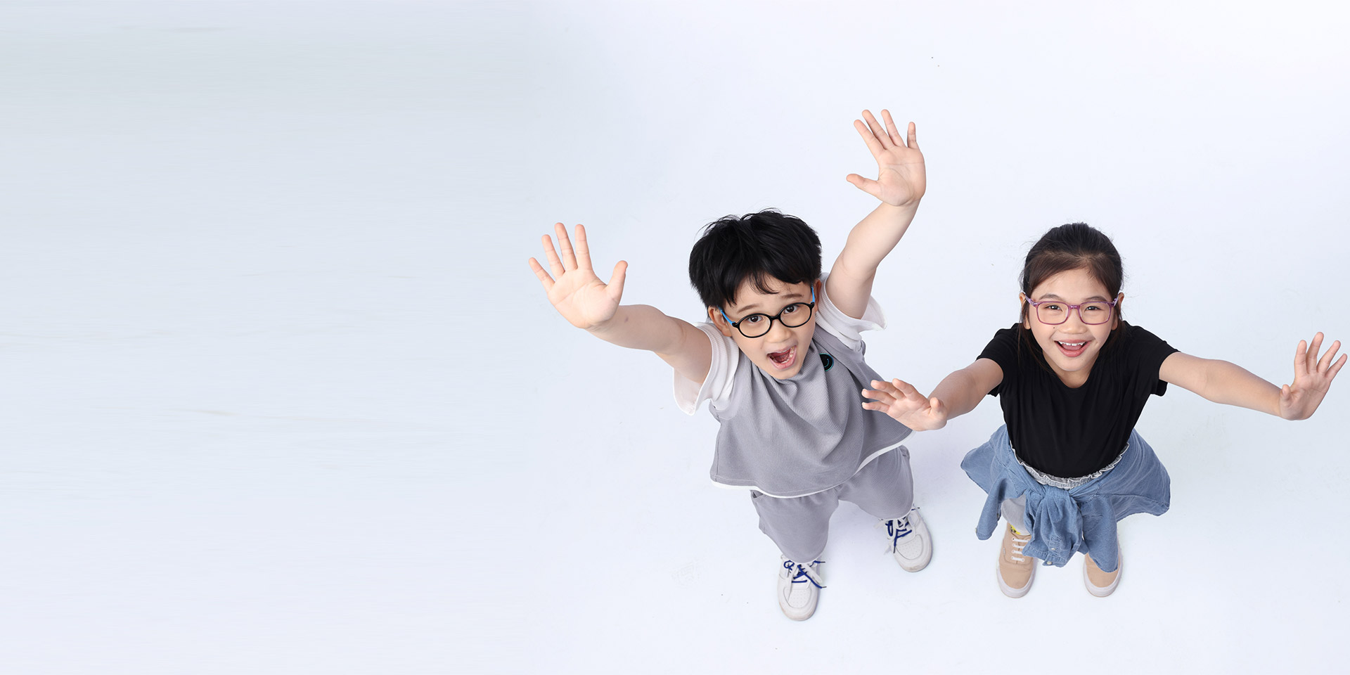 A boy and a girl, both wearing glasses, are looking up and putting their hands up in the air.