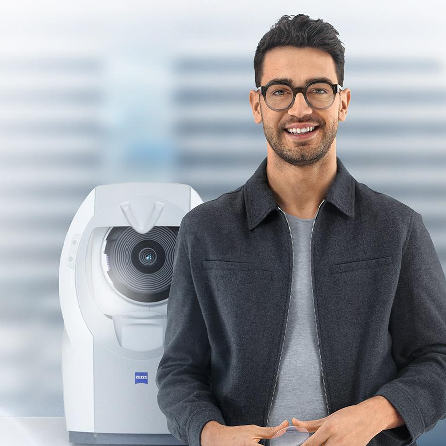 Young man with short brown hair smiling in camera wearing ZEISS lenses. In the background there is a ZEISS i.Profiler plus.