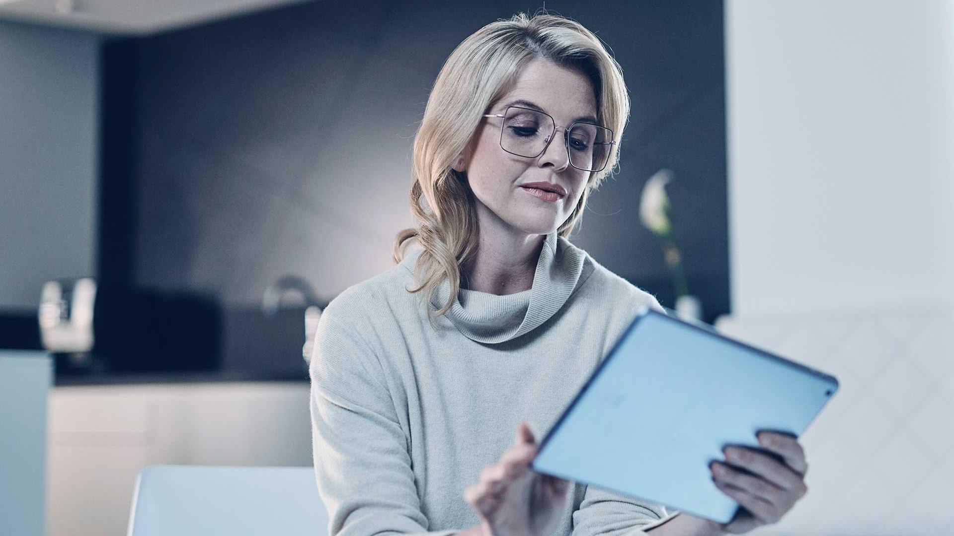 Blonde woman with glasses and a beige sweater holds a tablet and reads