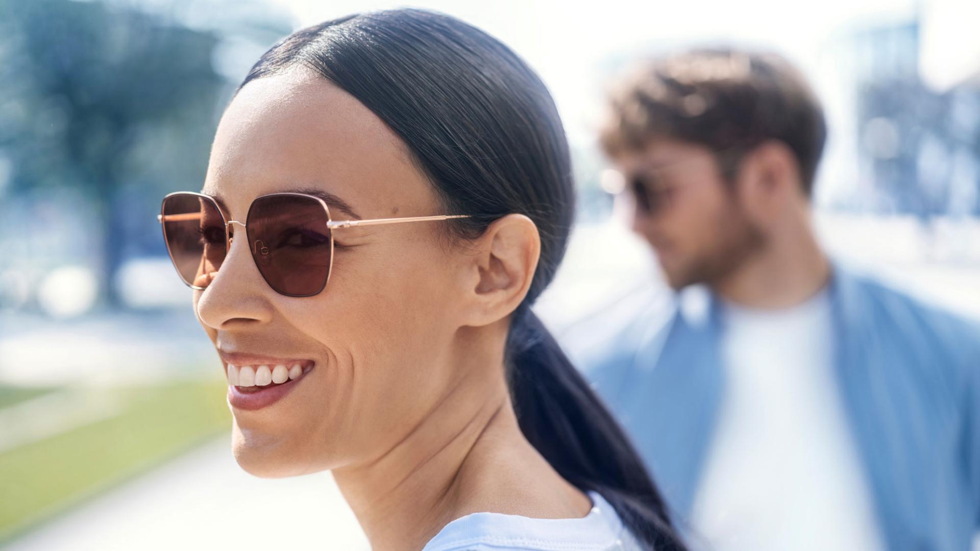 Front: smiling woman with sunglasses, background: a man with glasses