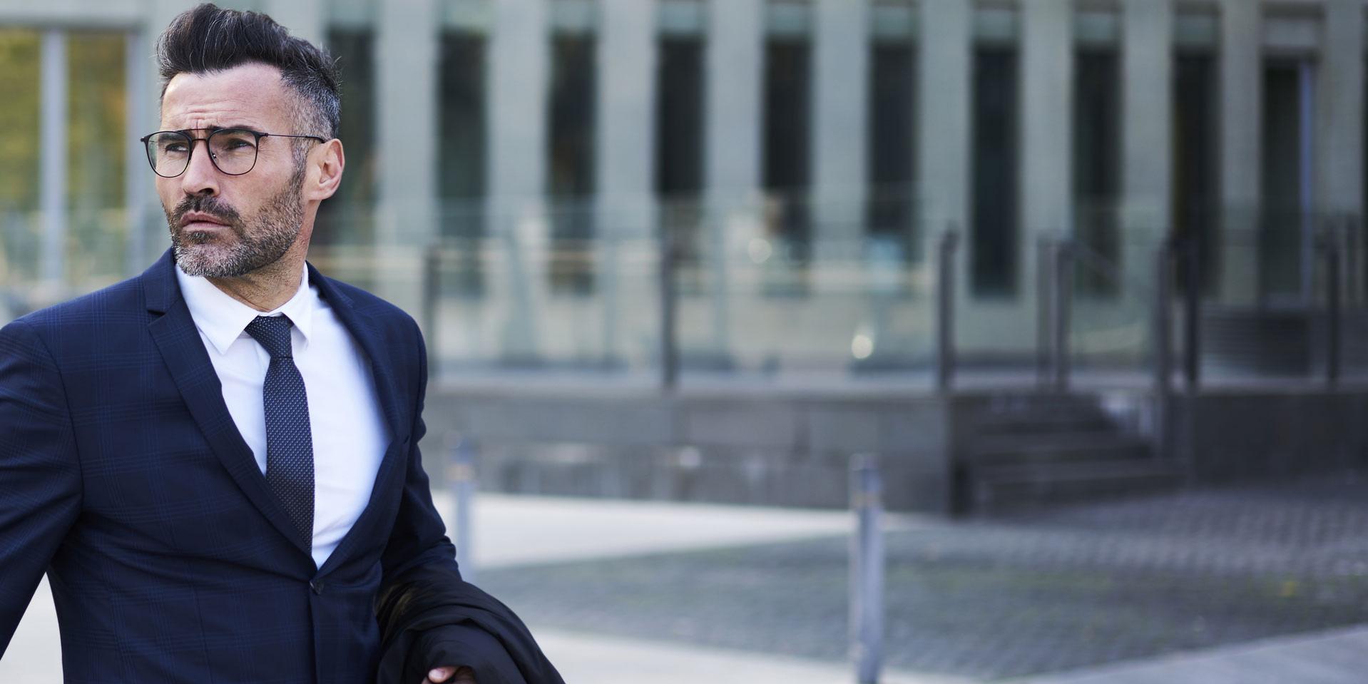 Confident handsome bearded male lawyer in trendy eyeglasses crossing street passing modern constructed architecture buildings with headquarters in business district of city hurrying for meeting.