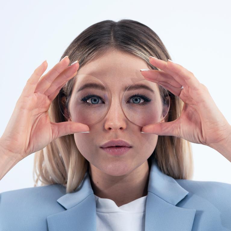 A young blonde woman holds lenses in front of her eyes to show good looks without any funny eye-effect due to ZEISS ClearView single vision lenses.
