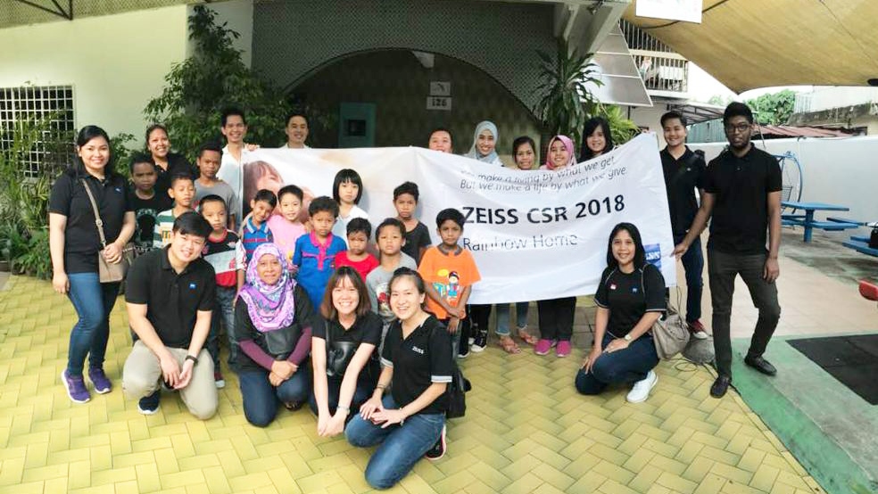 a group picture of ZEISS volunteers with the children of Rainbow Home orphanage, holding a banner with the headline "ZEISS CSR 2018, Rainbow Home" and a quote "we make a living by what we get but we make a life by what we give"