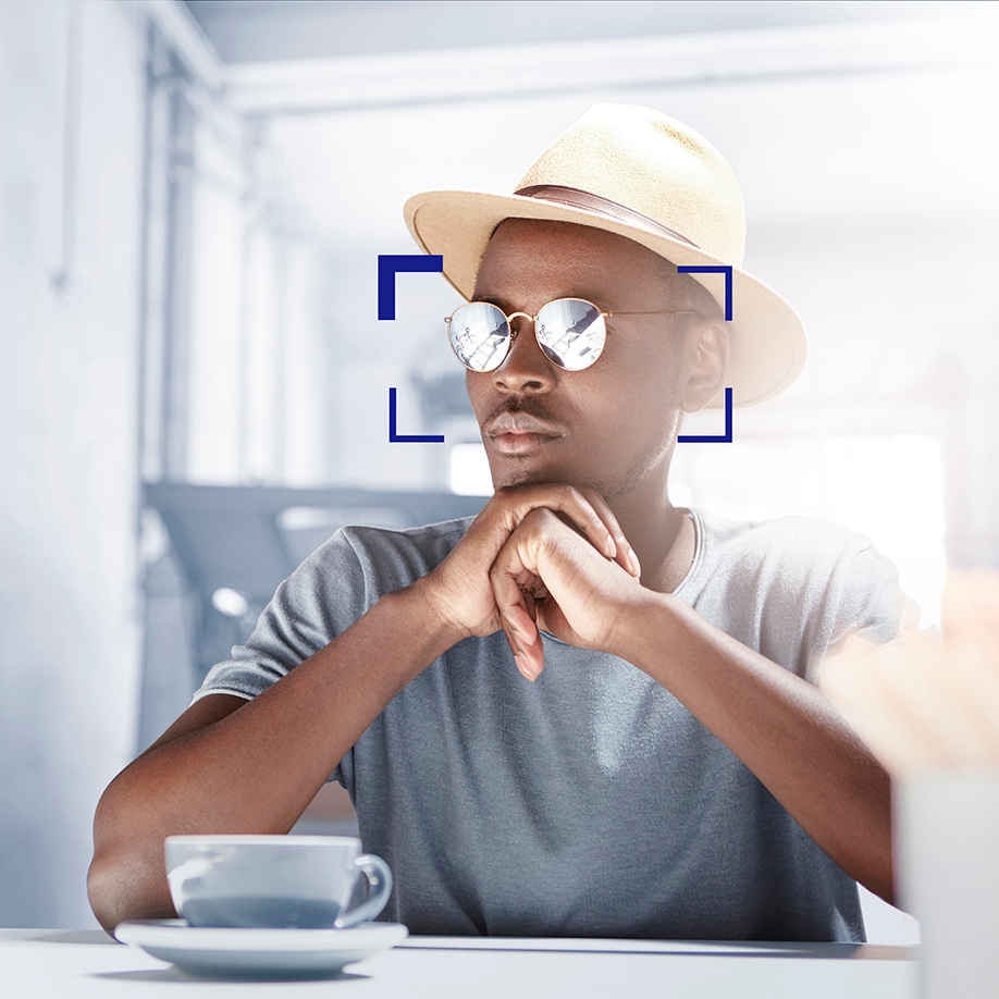 Indoor shot of attractive young Afro American man dressed in fashionable clothing and eyewear waiting for his girlfriend for lunch, sitting at cafe table with mug and cell phone, having pensive look